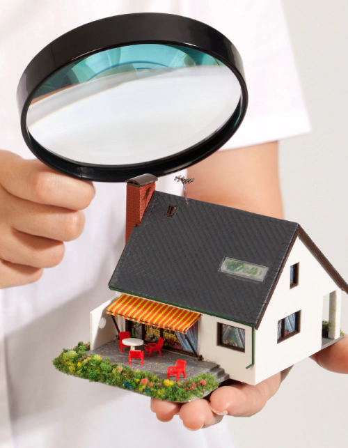 woman hand holds a magnifying glass over a miniature house
